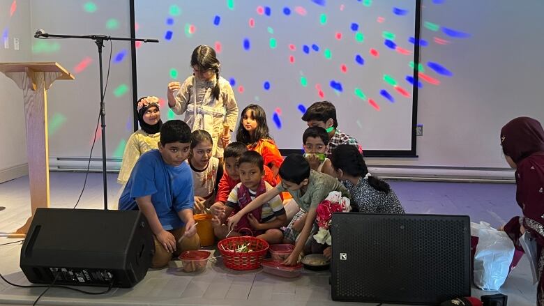 A group of children sit on a stage in various types of clothing with baskets in front of them on the stage.