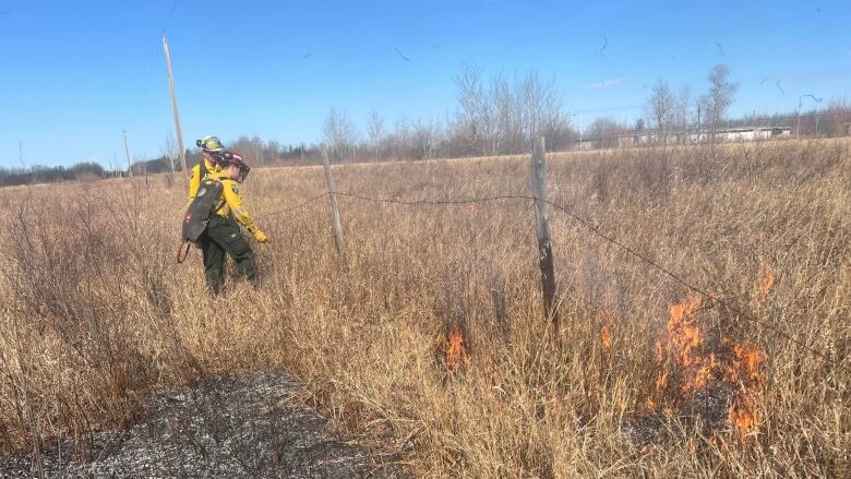 Two wildfire firefighters stading next to a wire fence, with a grass fire burning the foreground. 