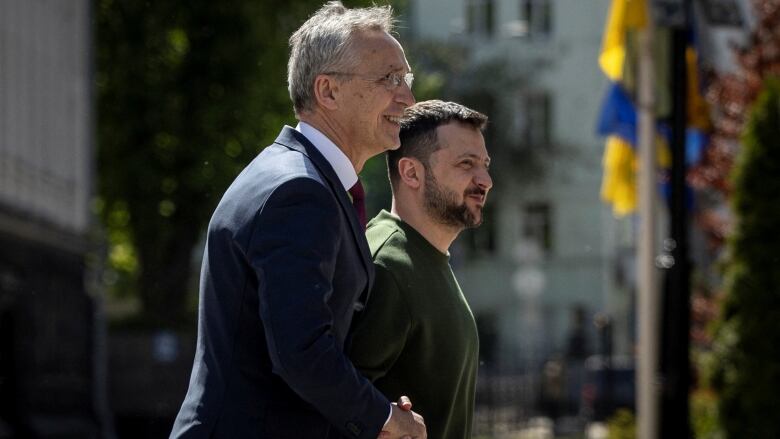 A bespectacled older man wearing a suit and tie and a younger bearded man in an army-coloured sweatshirt shake hands while outside.