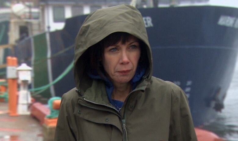 A woman wearing a green rain jacket holds a picture frame. Behind her is a harbour with boats tied to a wharf. 