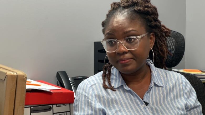 A woman sits in an office chair. She wears a blue and white striped shirt, clear glasses and silver hoop earrings. Her hair is braided away from her face.