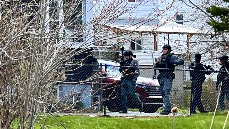 Police wearing body armour walk in the backyard of a home behind a fence, with a bush visible on the left hand side.