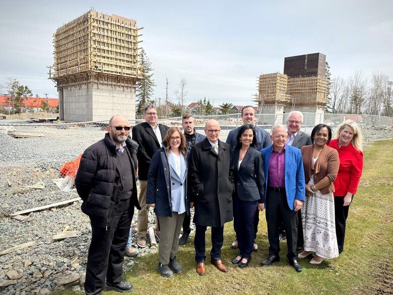 A group of people stand on green grass in front of a fenced-off construction zone containing gravel and several concrete and wooden structures in the back.