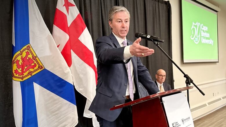 A man in a blue suit at a podium with two flags behind it gestures with his hand while another man looks on in the background.