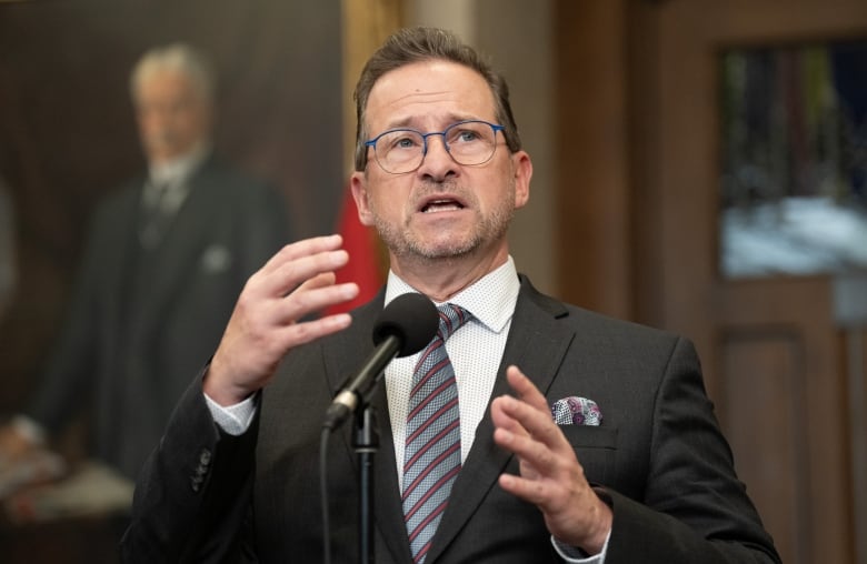 A man in a dark suit and striped tie speaks into a microphone. A portrait hangs in the background.