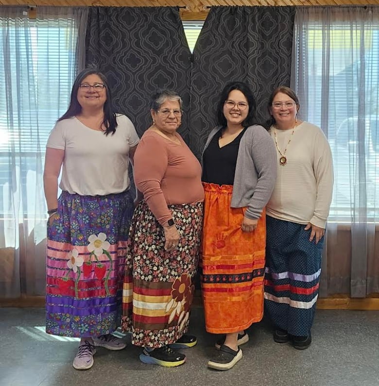 A group of four women standing in a line, each wearing a brightly coloured ribbon skirt.