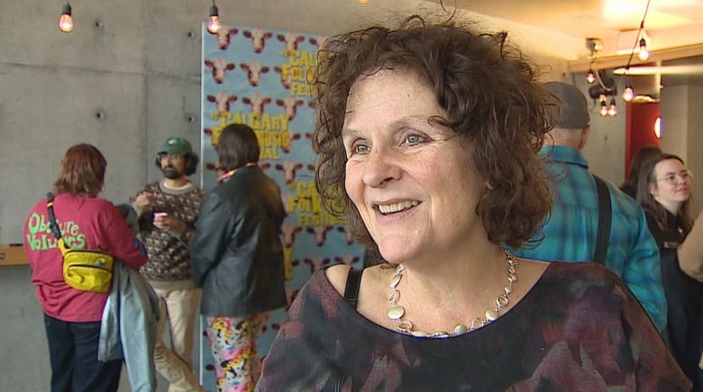 a woman with brown hair smiles and talks to a camera. there are people in the background.