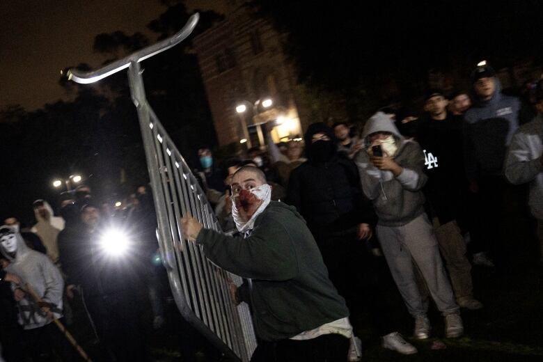 A man with a mask on his face lifts a metallic fence and appears ready to throw it.