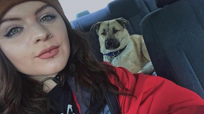 A selfie of a young woman with dark hair, sitting in a car, with a dog in the back seat behind her.