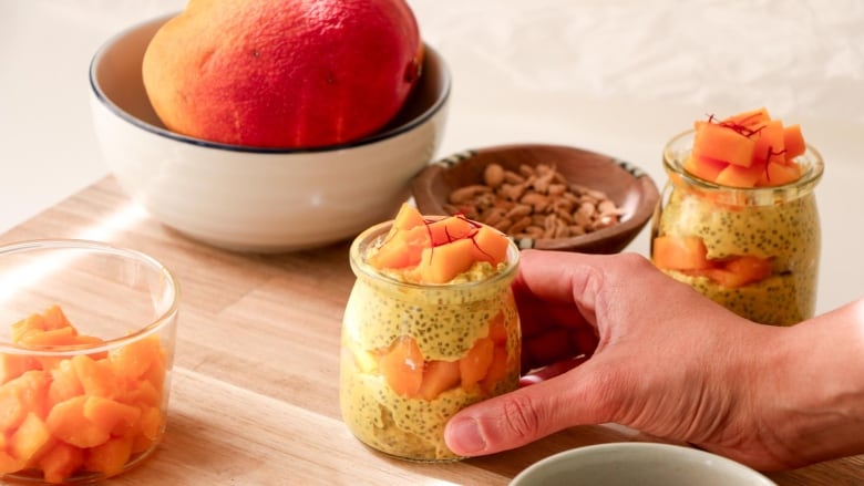 A hand grabbing a small glass pot of mango lassi chia pudding from a wooden cutting board. 