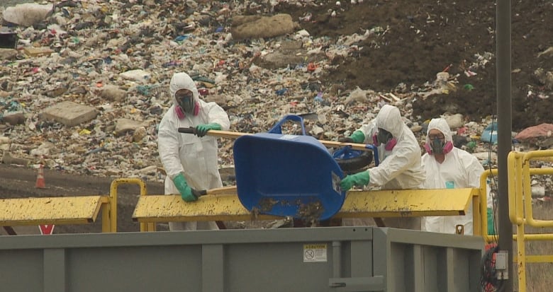 Three people in personal protective equipment including white suits, green gloves and respirators, dump a wheelbarrow's contents into what looks like a dumpster.