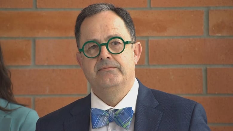 A man in a dark blue suit, blue and green bowtie and green-rimmed glasses. 