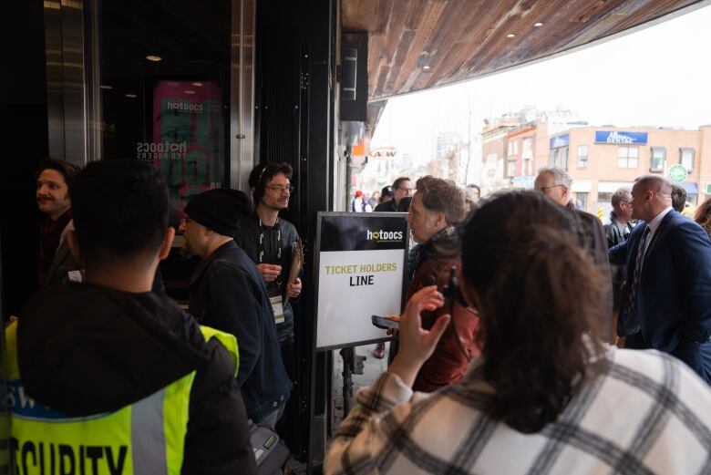 A crowd of people stand in front of theatre doors. A sign in front of them reads 'Ticket Holders line.'