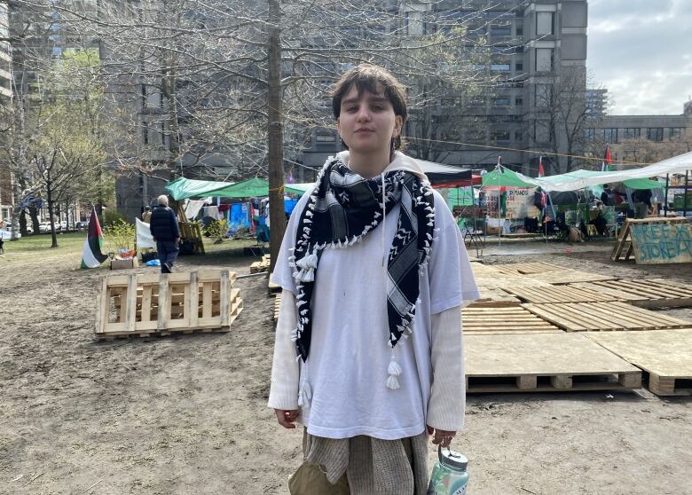 A young person with short hair and a keffiyeh scarf stands in front of an encampment