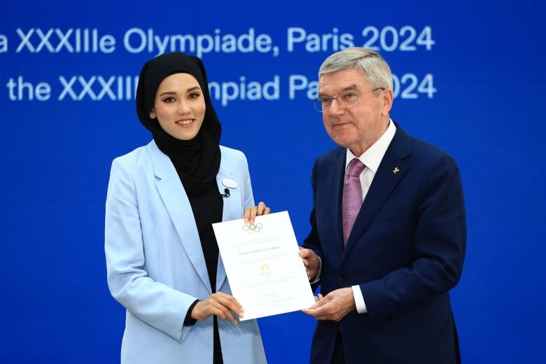 Two people pose for a photograph while holding a sheet of paper, in front of a blue background.