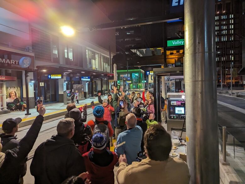 A crowd of people stand in the dark on an LRT platform cheering, one person raises comically large scissors in the air