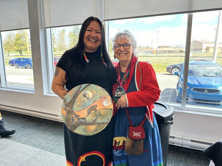 Two women wearing long skirts stand together. The woman on the right holds a painted drum.