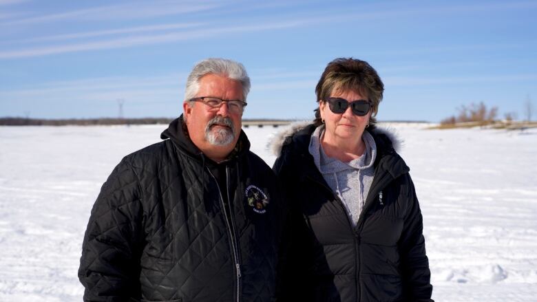 A man with short grey hair and glasses stands next to a woman with short brown hair and sunglasses as they stand on a frozen lake in the winter.