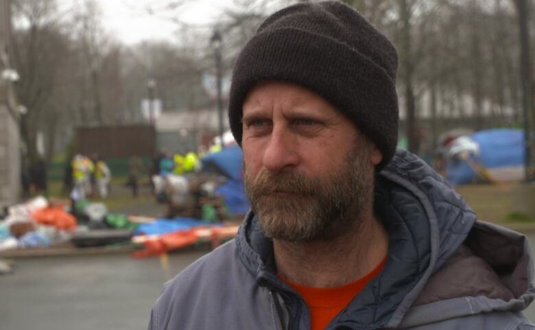 A man with a beard wearing a beanie and jacket stands in a parking lot. People and tents can be seen behind him.