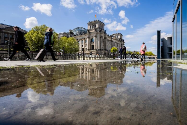 People walk outdoors on a sunny day past a historic building that is reflected by a large puddle.