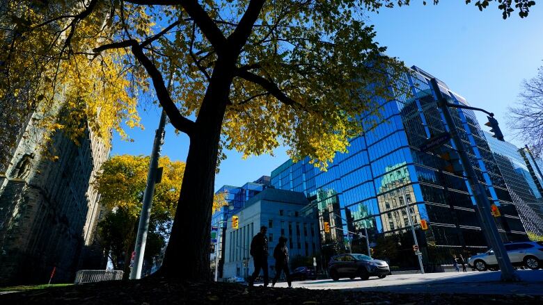 People in shadow walk past a glass building on a downtown street on a sunny day.