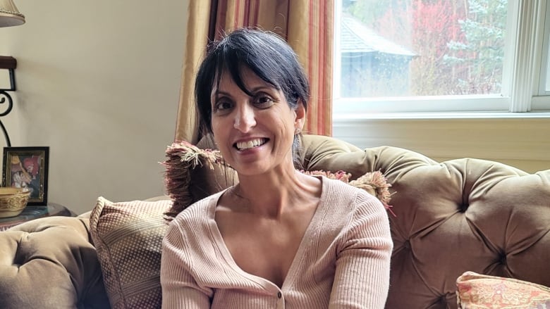 An adult woman of south asian descent with short dark hair smiles and sits on a couch inside a home living room.