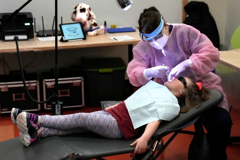 A dental hygienist examines a child's teeth. There are tables in the background.