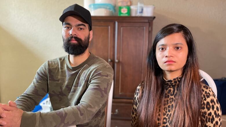 A man and woman sit at a kitchen table and look at the camera with solemn faces.    