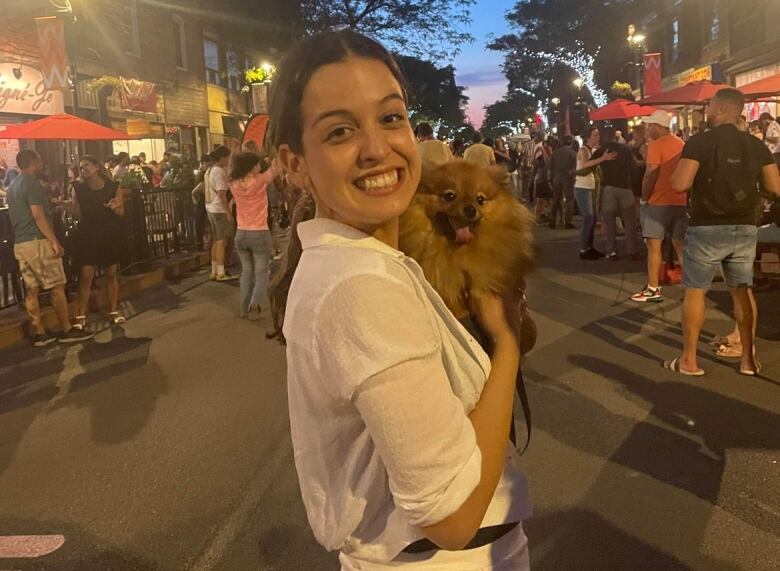 A woman stands on a street as dusk falls. She's smiling at the camera and holding a small, golden-haired dog.