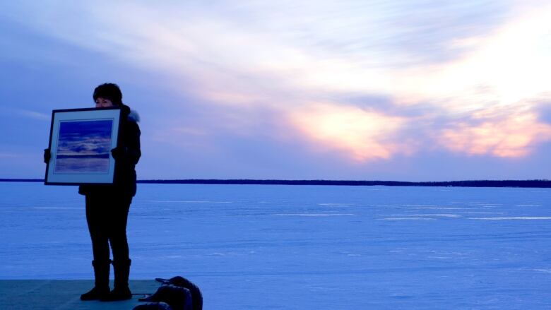 A woman holds up a painting of a purple and orange sunset while she stands on a dock overlooking a frozen lake at sunset with similar colours.