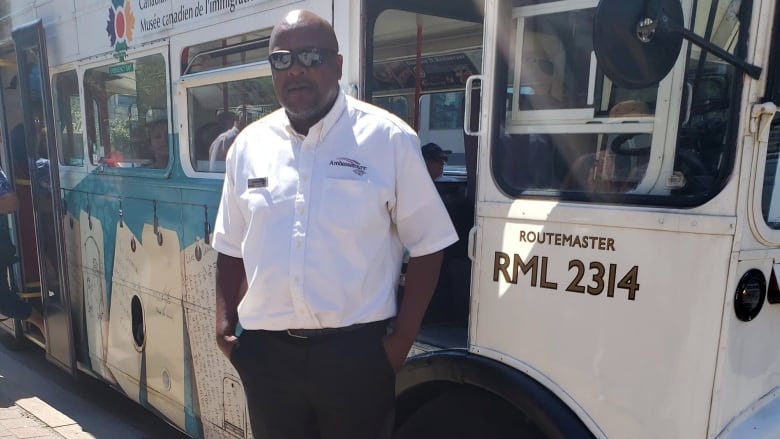 A Black man wearing a white shirt and black pants stands outside of a tour bus.