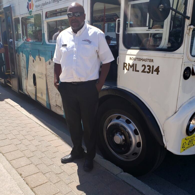 A Black man wearing a white shirt and black pants stands outside of a tour bus.