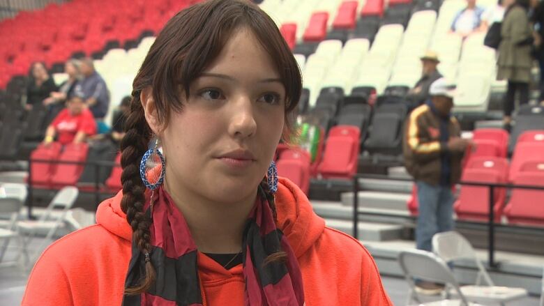 A girl in an orange hoodie stands in front of a row of chairs.