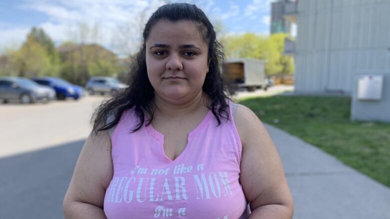 A woman in a pink t-shirt stands outside and looks at the camera.