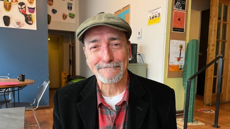 an older white man with grey facial hair wearing a red flannel shirt, black blazer and khaki green cap looking at the camera.