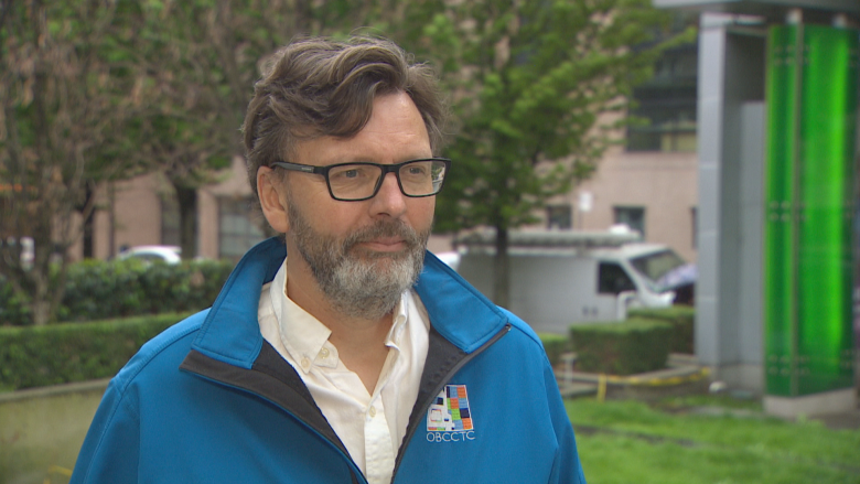 A man in glasses and a blue raincoat looks serious.