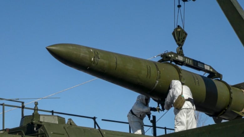 Two people covered from head to toe in baggy safety uniforms are shown near a large, cylindrical pointed weapon.