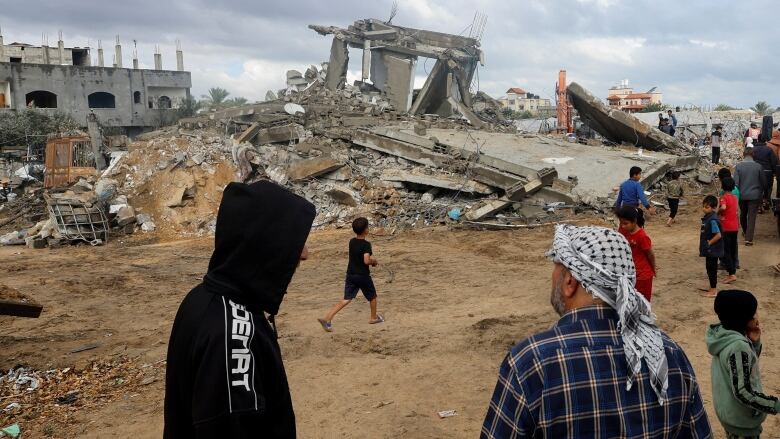 People in hooded sweaters stand across the street from the concrete rubble of a building.