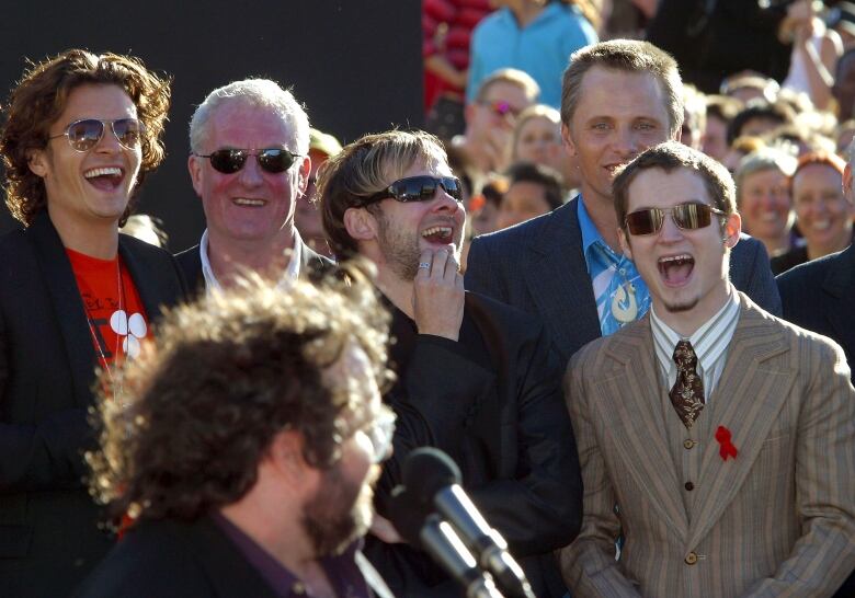 A smiling group of men wearing suits stand in front of a large crowd. 