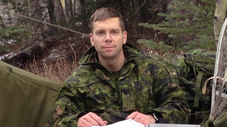 A man wearing military fatigues sit in the woods with a clipboard and snowshoes. 