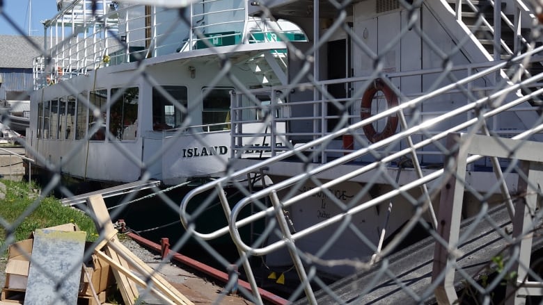 Two large, white boats with life preservers hanging from them are shown behind a chain-link fence.