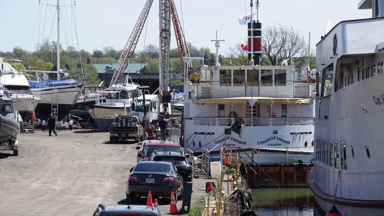 A large white boat is shown floating at a dock behind an even bigger, white cruise ship. Workers can be seen walking around them and they're surrounded by cranes, equipment and parked cars.