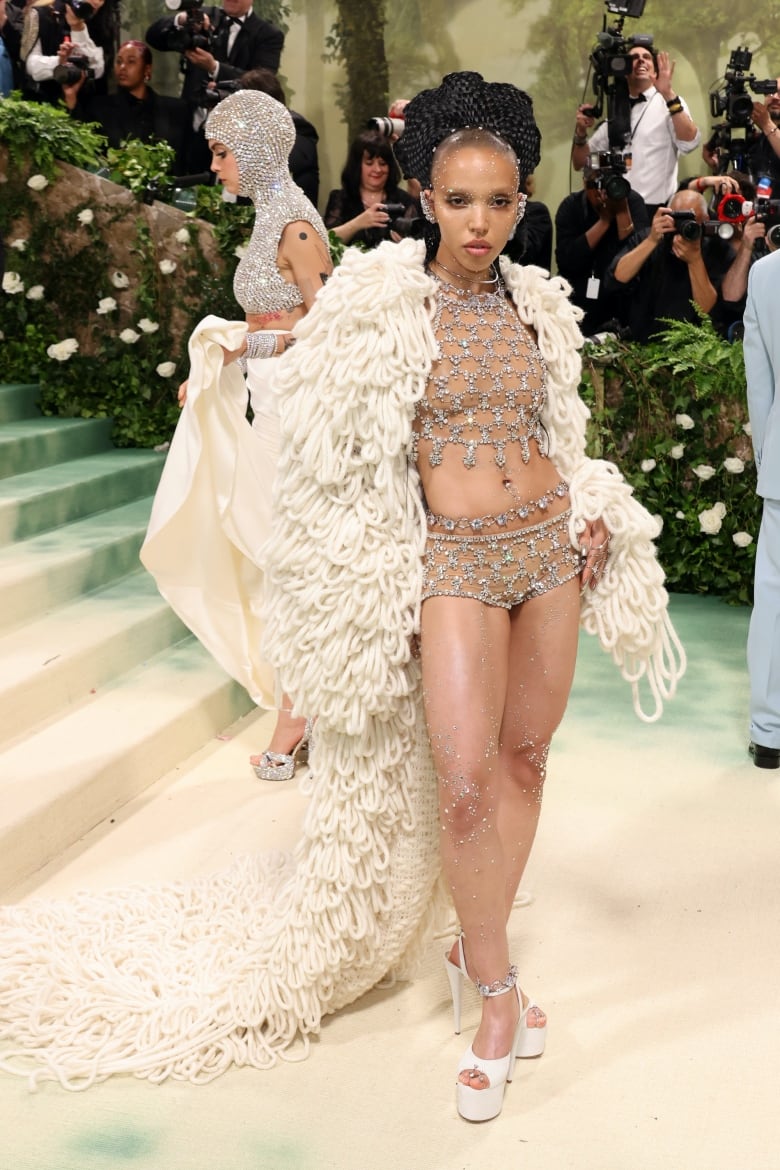 FKA twigs on the Met Gala red carpet wearing a cream cape with knitted loops and a 2-piece diamond-embellished top and bottoms. 