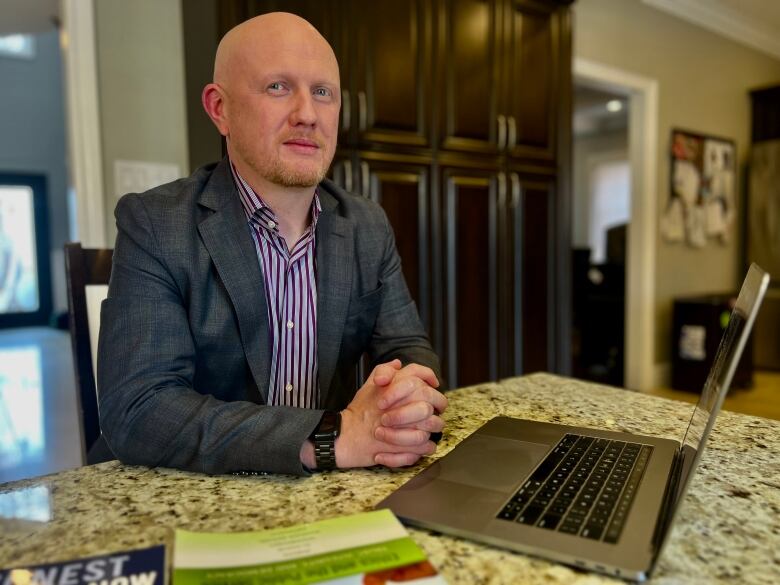 A man in a grey suit with a purple pinstripe shirt sits at a kitchen island with a laptop open. He looks at the camera. 