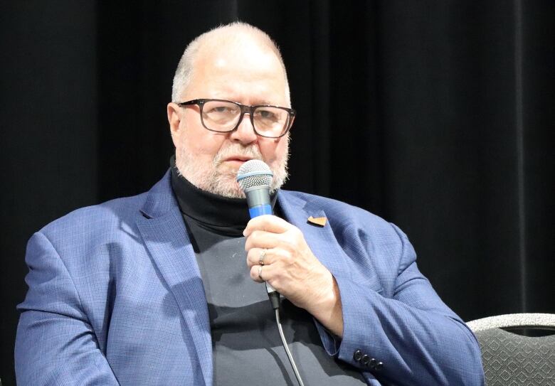 A balding man with a blue suit, charcoal turtleneck sweater and glasses holds a microphone in his hand while speaking.