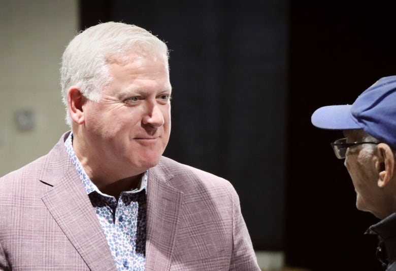 A man with white hair and a mauve jacket and print shirt smiles.