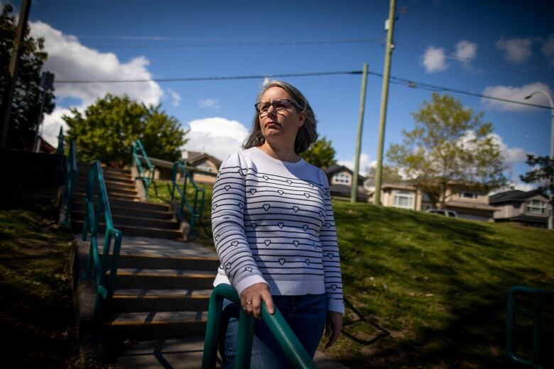 A woman, peering off to the left, pauses to hold a handrail as she walks down a set of stairs outdoors next to a high school on a sunny spring day.