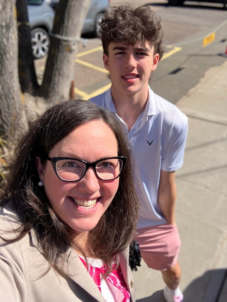 A white lady with brown hair, wearing glasses. Her son with a white complexion and curly brown hair stands behind her.