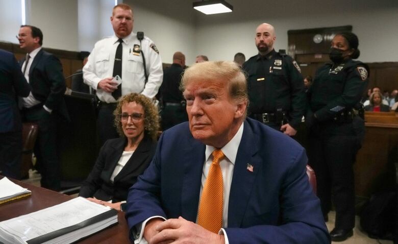 A man in a blue suit with an orange tie sits with his hands folded on the table in front of him.