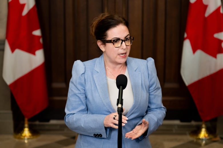 A woman in a blue blazer speaks into a microhpone. Two Canadian flags are draped in the background.